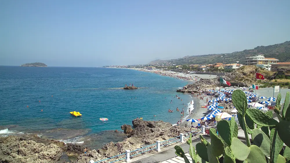 Spiaggia con persone che nuotano e giocano in acqua, con un paesaggio costiero e cielo blu sullo sfondo.
