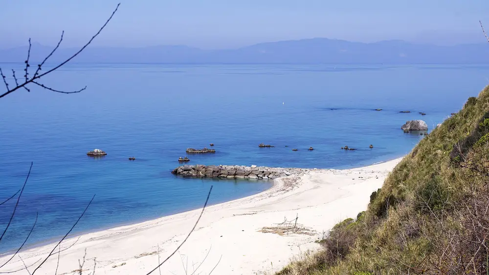 Spiaggia con sabbia bianca e acqua blu profonda, con rocce sparse nel mare e montagne sullo sfondo.