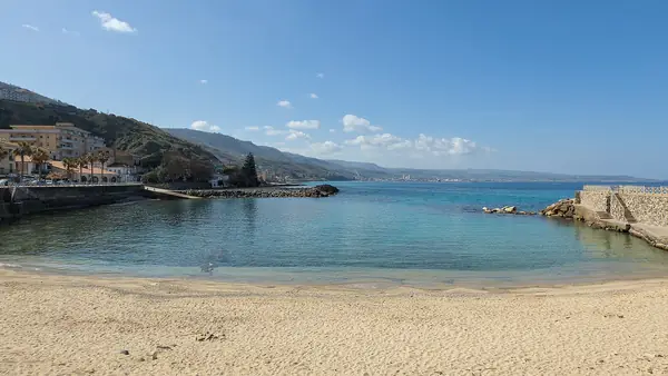 Spiaggia con sabbia dorata e acqua blu profonda, con una vista panoramica sul mare e sulla città sullo sfondo.