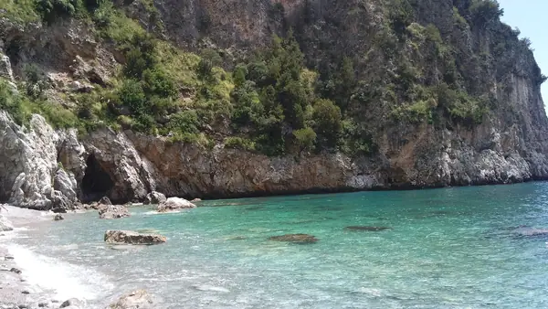 Spiaggia rocciosa con acqua cristallina e montagne verdi sullo sfondo.