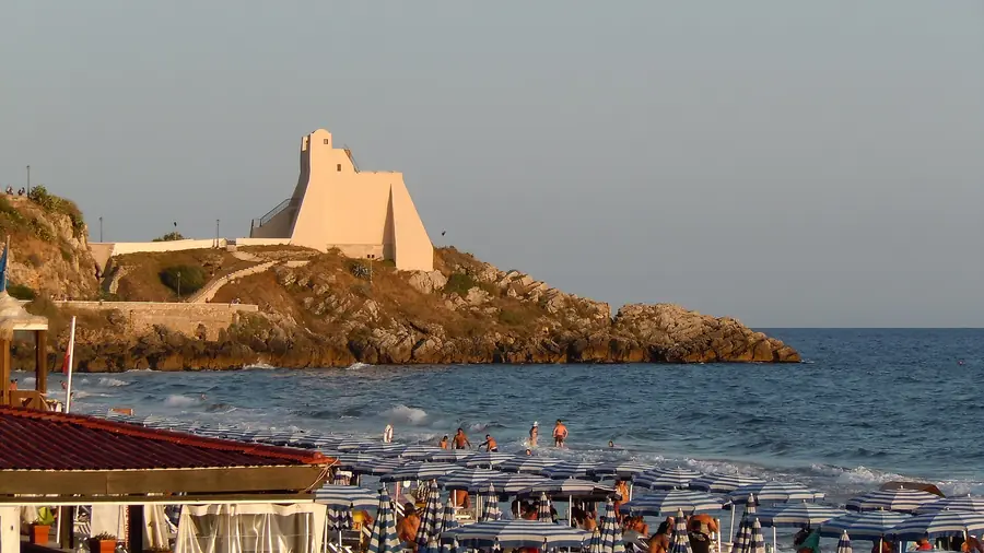 Spiaggia con ombrelloni a strisce blu e bianche, persone che si godono il mare e una torre sulla riva rocciosa.