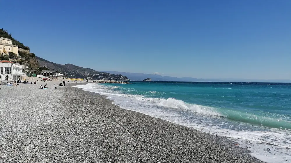 Spiaggia rocciosa con acqua turchese e cielo blu, persone che si rilassano sulla sabbia.