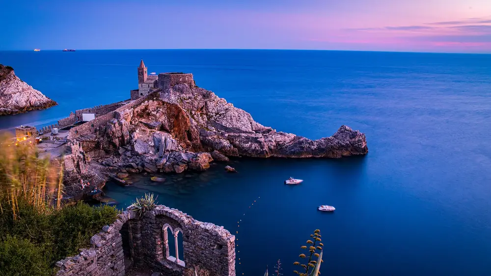 Un castello medievale costruito su una scogliera sopra un'isola con due barche in acqua e un tramonto blu sullo sfondo.