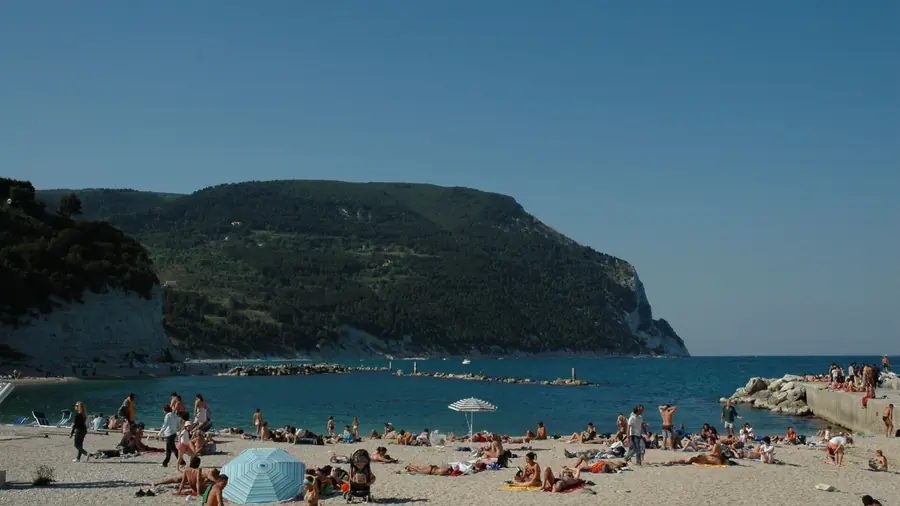 Spiaggia di Sirolo con molti turisti che si rilassano sulla sabbia e sotto gli ombrelloni, con il mare blu profondo e la montagna verde sullo sfondo.