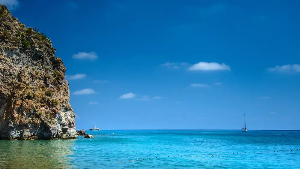 Spiaggia con acqua cristallina e cielo blu, con una barca e una scogliera sullo sfondo.